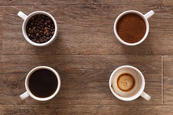 Variedad de tazas de café y granos de café en la vieja mesa de madera. Cuatro tazas de café, fases de bebida - frijol, taza molida y vacía . — Foto de Stock