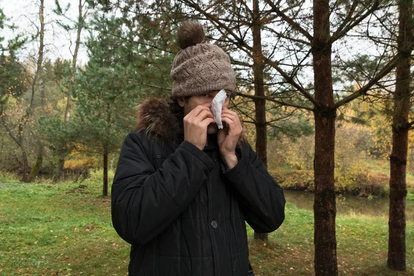 Young man wearing jacket suffering from cold and holding handkerchief on nose. Male in knitted hat standing in the wood blowing his nose. Autumn illness and flu season concept.