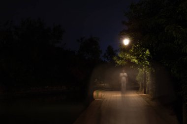 Double exposure night scene of person walking dark street illuminated with streetlights. The receding male silhouettes on the road in the park. Human figure in motion blur going along the city river clipart