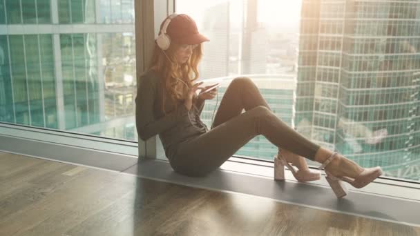 Linda chica sentada por la ventana escuchando música con auriculares y enviando sms usando el móvil — Vídeos de Stock