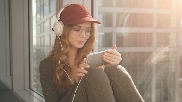 Χαριτωμένο κορίτσι που κάθεται δίπλα σε παράθυρο, να ακούτε μουσική με ακουστικά χρησιμοποιώντας το κινητό — Αρχείο Βίντεο