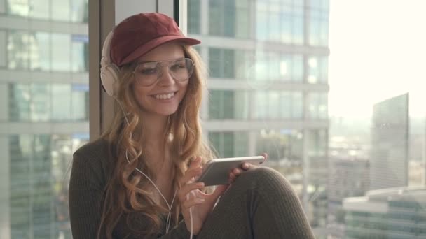 Chica bonita sentada por la ventana escuchando música con auriculares y sonriendo feliz mirando a la cámara — Vídeo de stock