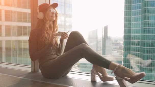 Menina bonito sentado pela janela ouvir música com fones de ouvido e enviando sms usando celular — Vídeo de Stock