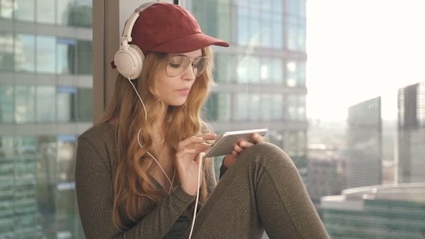 Chica sonriente sentada por la ventana escuchando música con auriculares usando el móvil — Vídeos de Stock