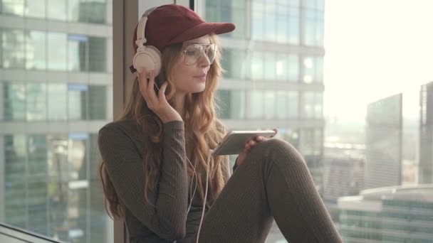 Chica sonriente sentada por la ventana escuchando música con auriculares usando el móvil — Vídeos de Stock