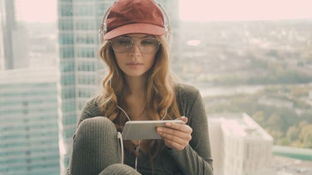 Νεαρή γυναίκα που κάθεται κοντά σε παράθυρο, να ακούτε μουσική με ακουστικά χρησιμοποιώντας το κινητό — Αρχείο Βίντεο