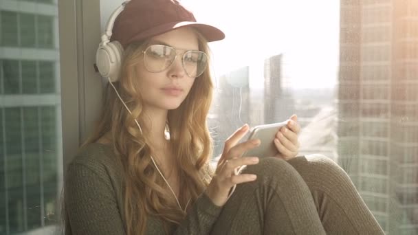 Menina muito sorridente sentado à janela ouvir música com fones de ouvido usando celular — Vídeo de Stock