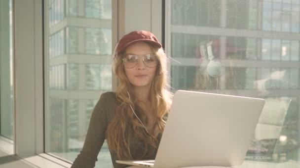 Hezká dívka sedí u okna s notebookem a poslouchat hudbu na pozadí města — Stock video