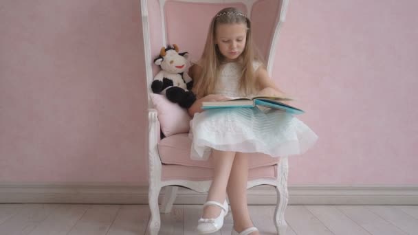 Lovely little girl with long hair in white dress sit on pink chair and reading book — Stock Video