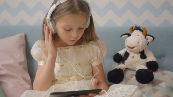 Linda niña con auriculares está escuchando música usando una tableta, mientras está sentada en el sofá en casa — Vídeos de Stock