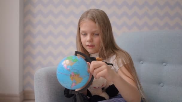 Симпатична маленька дівчинка тримає і обертає маленький глобус, сидячи на дивані — стокове відео