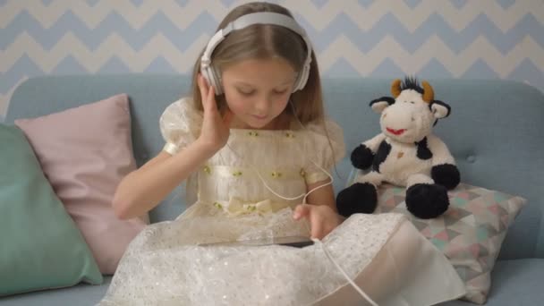 Linda niña con auriculares está escuchando música usando una tableta, mientras está sentada en el sofá en casa — Vídeo de stock