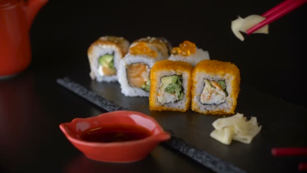 Mano con palillos rojos tomando jengibre y rollo de sushi de plato oscuro — Vídeos de Stock