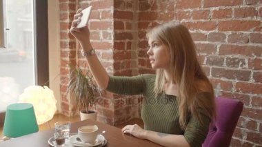 Güzel genç kadın bir akıllı telefon kullanarak ve kafede istirahat ederken gülümseyen selfie yapıyor