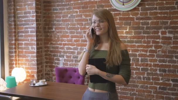 Νέοι χαριτωμένο κορίτσι με τα γυαλιά, στέκεται σε δωμάτιο κοντά σε τοίχο από τούβλα και να μιλάει στο κινητό, κρατώντας τον υπολογιστή tablet στο χέρι. — Αρχείο Βίντεο