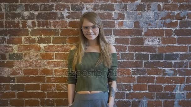 Beautiful young girl with tattoo is looking at camera and smiling while standing against red brick wall — Stock Video