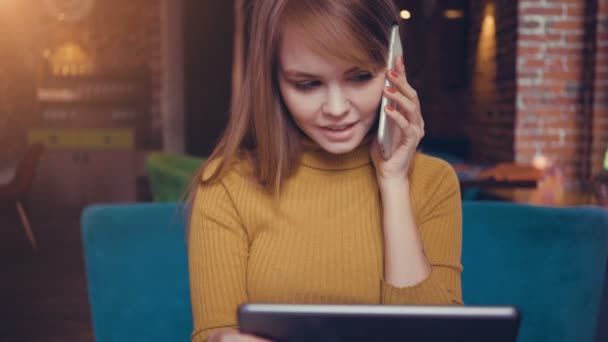 Junge süße Frau telefoniert mit Handy, während sie mit digitalem Tablet im Café sitzt — Stockvideo