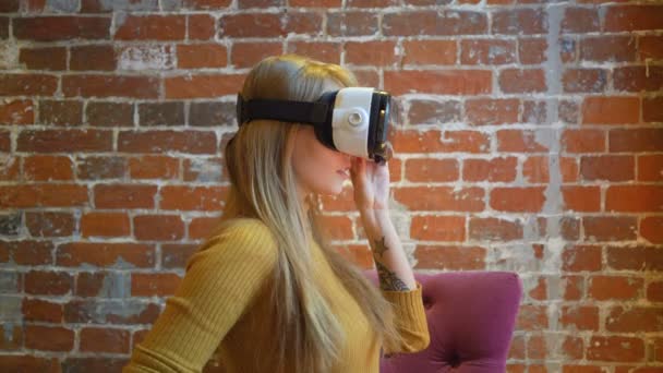 Привлекательная молодая женщина в наушниках VR жестикулирует и улыбается, сидя дома — стоковое видео
