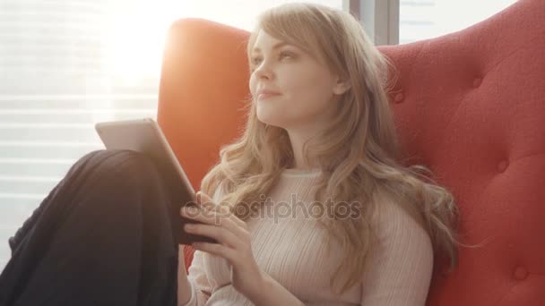 Jeune femme à la maison assise sur un fauteuil rouge devant la fenêtre et travaillant avec une tablette — Video