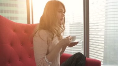 Genç kadın evde modern kırmızı sandalye oturma odasında rahatlatıcı ve kahve veya çay içme pencerenin önünde oturup.....