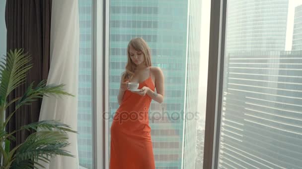 Fiatal nő visel piros hálóruhaként csésze teát iszik, és az ablak mellett állandó — Stock videók