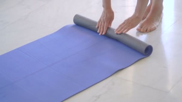 Женщина складывает синий коврик для йоги или фитнеса после тренировки дома в гостиной . — стоковое видео