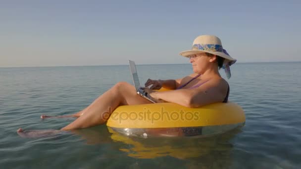 Женщина средних лет, работающая на ноутбуке в оранжевом надувном кольце в отпуске — стоковое видео
