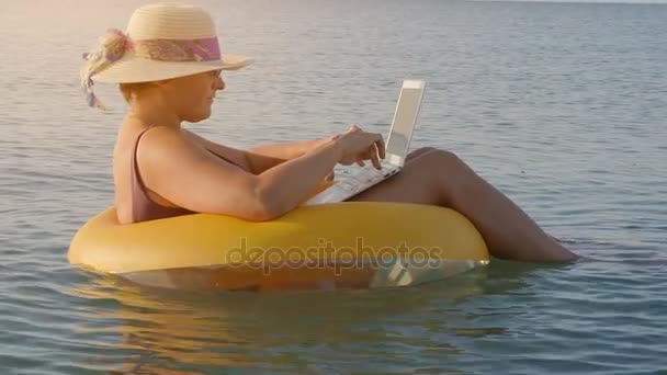 Female freelancer works on a laptop sitting in an inflatable ring in the sea, — Stock Video