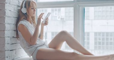 Pencere kenarında oturan ve kulaklık ile smartphone kullanarak seksi kadın
