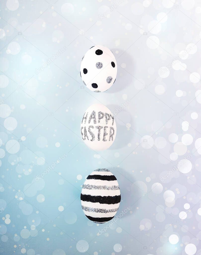 Composition of glitter inscription Happy Easter on a white egg and shining bokeh on gradient background. Eggs with black, silver lines and polka dots. Flat lay, vertical top view, close up, copy space