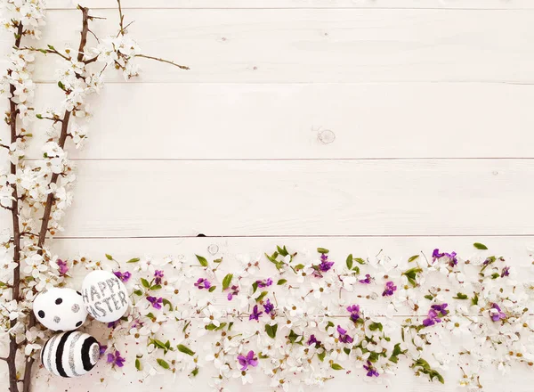 Composition of spring flowers, blooming tree branches and eggs with black, silver lines and polka dots on wooden background. Glitter inscription Happy Easter. Flat lay, top view, close up, copy space