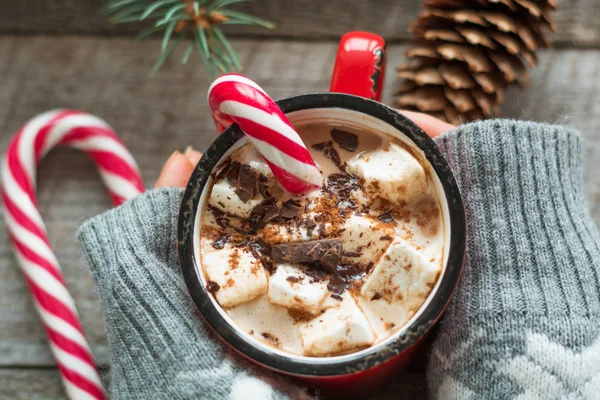 Christmas hot beverage hold hand in sweater. Hot chocolate with marshmallows and candy cane on the wooden background. New Year. Holiday card. Rustic style. Top view and Close up.