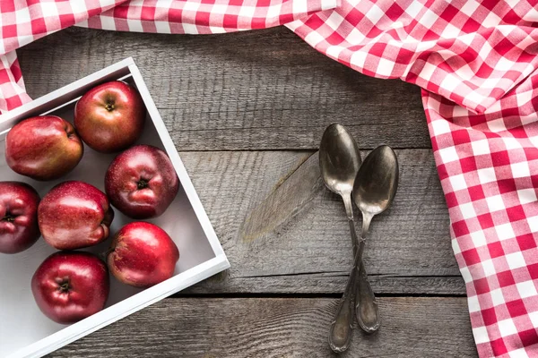 Manzanas rojas maduras en caja de abedul en tablero de madera con servilleta a cuadros roja alrededor, y cucharas y espacio de copia para su receta . — Foto de Stock