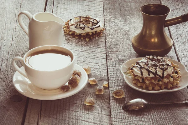 Белая чашка кофе с молоком, кувшин белого молока, коричневый сахар и домашние вафли со взбитыми сливками с декором сверху. Вкусный завтрак. Винтажный тонированный . — стоковое фото