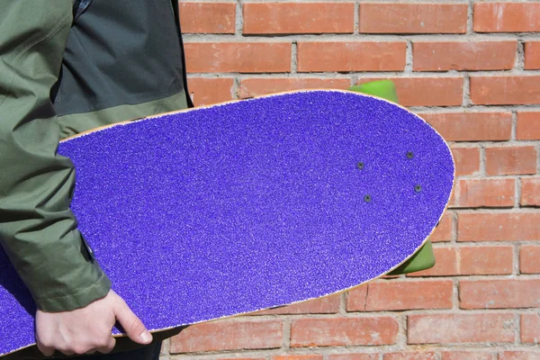 Teen držet fialové longboard cihlové zdi. Malá hloubka ostrosti. Detailní záběr. — Stock fotografie