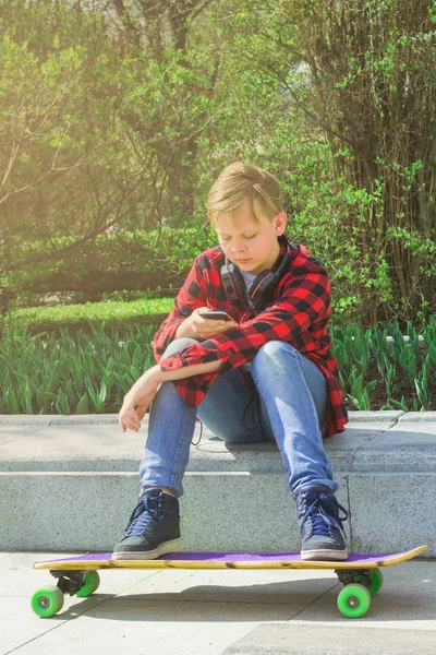Retrato al aire libre de adolescente moderno con camisa a cuadros roja con teléfono móvil y auriculares. Adolescente navegando por la red mientras está sentado en el parque, buscando gadget . — Foto de Stock
