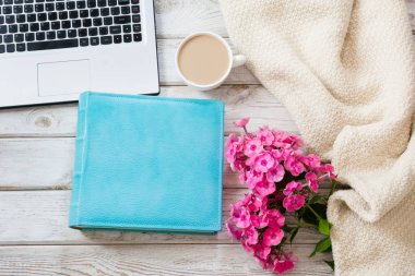 Kadın çalışma alanı ile bir dizüstü bilgisayar, fincan kahve, mavi defter, rahat kazak ve hafif ahşap zemin üzerine pembe çiçekler. Kopya alanı ile üstten görünüm kompozisyon.