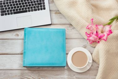 Kadın çalışma alanı ile bir dizüstü bilgisayar, fincan kahve, mavi defter, rahat kazak ve hafif ahşap zemin üzerine pembe çiçekler. Kopya alanı ile üstten görünüm kompozisyon.