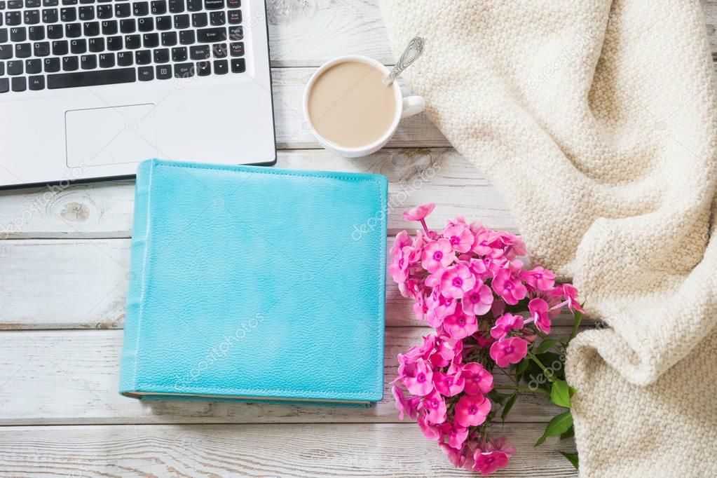 Female workplace with a laptop, cup of coffee, blue album, cozy sweater and pink flowers on light wooden background.Top view composition with copy space.