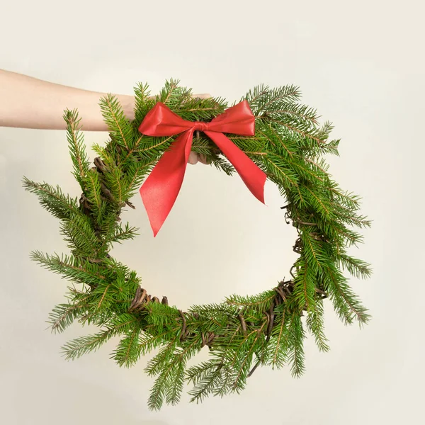 Corona natural fresca de Navidad de rama de abeto con lazo rojo en manos femeninas sobre fondo blanco. Imagen cuadrada . — Foto de Stock