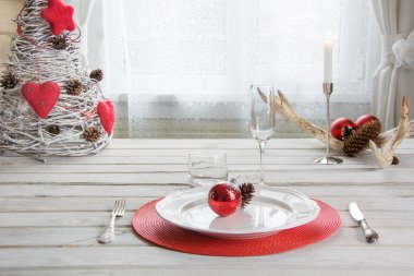 Noel sofra beyaz dishware, çatal bıçak takımı, gümüş ve kırmızı süslemeleri ligth iç pencere yakınındaki beyaz ahşap tahta üzerinde yer. Dekor olarak Noel şamdan.