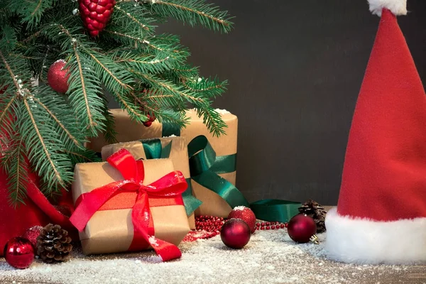 Fondo de vacaciones de Navidad. Regalos con una cinta roja, sombrero de Santa Claus y decoración bajo un árbol de Navidad en una tabla de madera. Copiar espacio en pizarra . — Foto de Stock