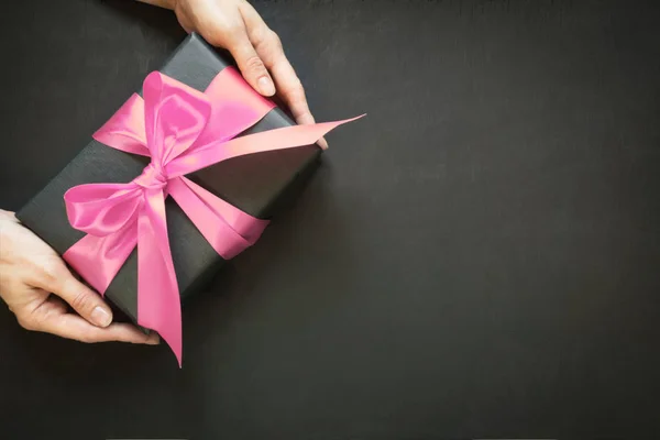 Geschenkverpackung in schwarzem Papier mit rosa Satinband in weiblicher Hand auf schwarzer Oberfläche. Kopierraum. Blick von oben. — Stockfoto