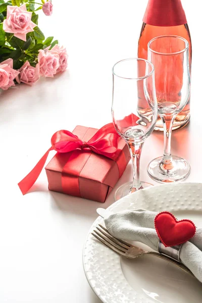 Cena di San Valentino con tavolo apparecchiato con regalo rosso, bicchiere per champagne, bottiglia di champagne, cuori con argenteria su sfondo bianco. Chiudete. Biglietto di San Valentino . — Foto Stock