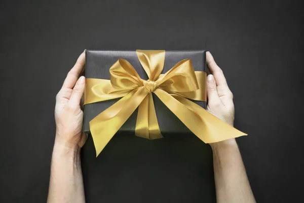 Geschenkverpackung in schwarzem Papier mit goldenem Band in weiblicher Hand auf schwarzer Oberfläche. Ansicht von oben. — Stockfoto