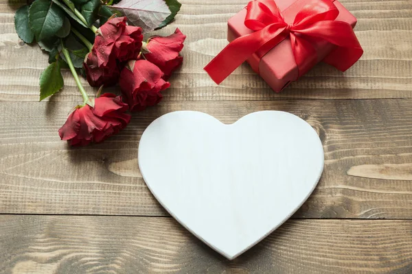 Μπουκέτο με κόκκινα τριαντάφυλλα και δώρο με κόκκινη κορδέλα και την καρδιά σε ξύλινη σανίδα. Κάρτα του Αγίου Βαλεντίνου. Χώρο αντίγραφο. — Φωτογραφία Αρχείου