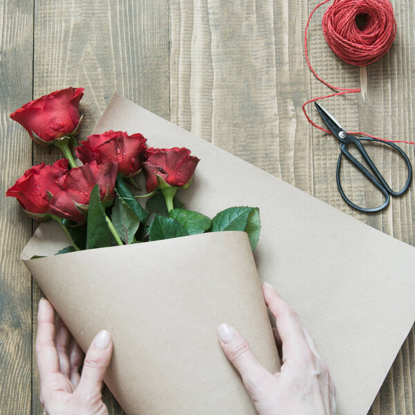 Флорист делает букет из красных роз, завернутый в крафтовую бумагу на деревянном столе. Вид сверху. Сельский стиль
.