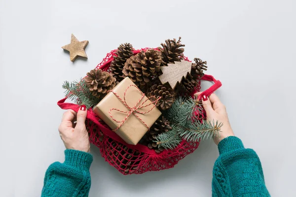 Woman hands holding mesh bag with Christmas zero waste decor, sustainable gift, cones and fir tree branches for handmade design.