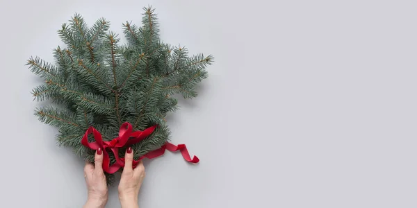 Mano de mujer sosteniendo ramas de árbol de Navidad con cinta roja sobre gris. Tarjeta de felicitación de Navidad . — Foto de Stock