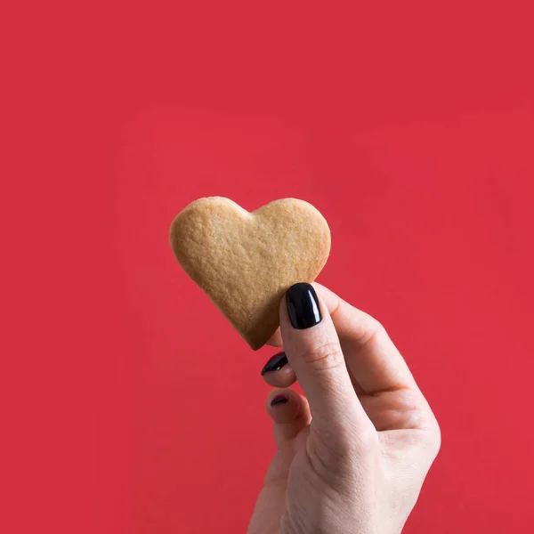 Biscotti a forma di cuore durante il Giorno di San Valentino in mano femminile su rosso. Concetto . — Foto Stock